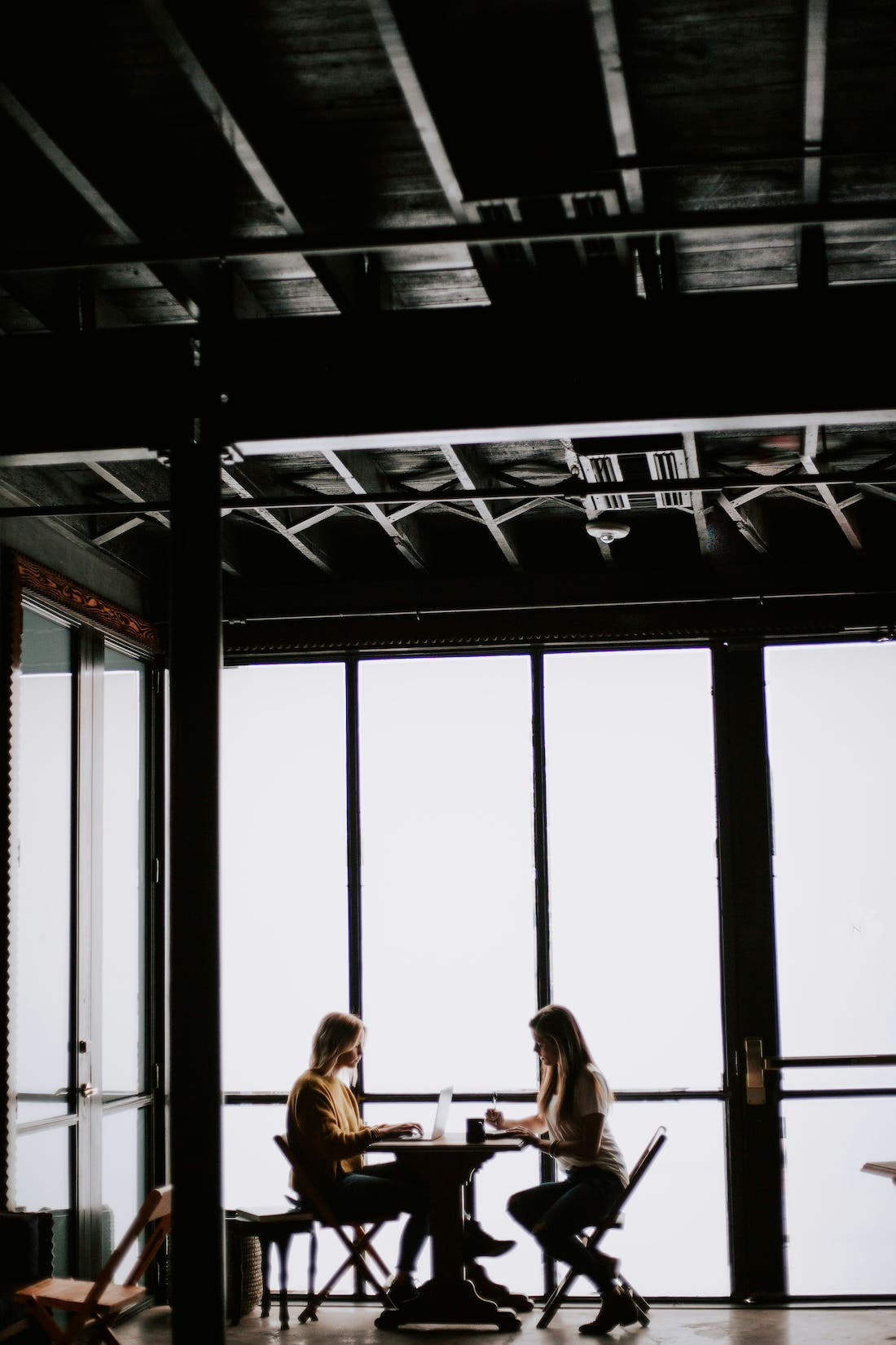 A medium shot photograph of two people sitting at a table in a dimly lit room, silhouetted against a bright window. The room has a high ceiling with exposed beams and a dark, industrial aesthetic. The two people are seated facing each other, engaged in conversation or working on something at the table. They appear to be indoors, possibly in a cafe or workspace, with large windows providing a stark contrast between the dark interior and the bright outside light. The image emphasizes the silhouettes of the people and the architectural details of the room.