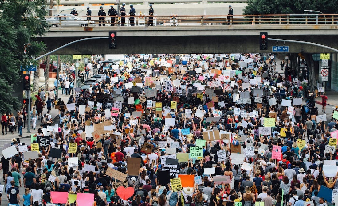 A high-angle, medium shot photograph captures a large crowd of people marching down a city street. The crowd is dense, and many individuals are holding up signs, some of which are partially visible. A bridge or overpass crosses the street overhead, with a line of police officers standing on it, observing the crowd. Street signs for "4th St" are visible at an intersection. The overall scene suggests a protest or demonstration.