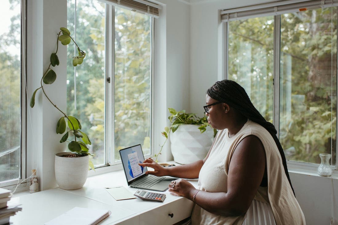 A medium shot of a person with dark braided hair wearing glasses and a light-colored dress, sitting at a white desk and working on a laptop. The person is indoors, positioned between two large windows that provide a view of trees. A Golden Pothos plant is visible in a white pot on the left side of the desk, and another plant is in a white pot on a windowsill to the right. A calculator and other items are also on the desk. The scene is brightly lit by natural light.