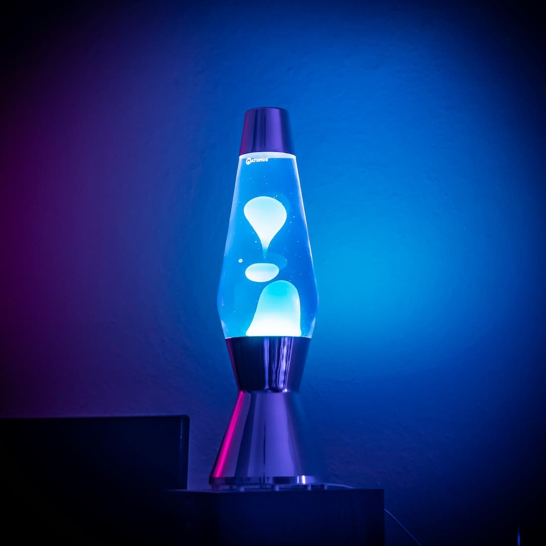 The image shows a blue lava lamp against a gradient background transitioning from purple to blue. The lamp has a silver base and top, and the blue liquid inside contains white, blob-like forms. The lamp is lit up, creating a glowing effect.