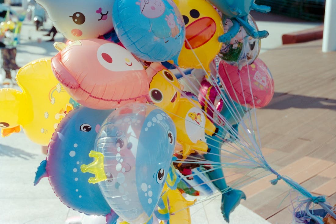 A large cluster of colorful foil balloons shaped like various cartoon characters and animals. Notable balloons include a light blue whale or dolphin, a yellow duck, a pink owl, a blue balloon with Peppa Pig, and several other light blue balloons with faces, some featuring transparent sections with smaller balloons inside. The balloons are tied together with numerous thin strings and are shown outdoors on what appears to be a wooden deck or boardwalk, with blurry figures in the background.